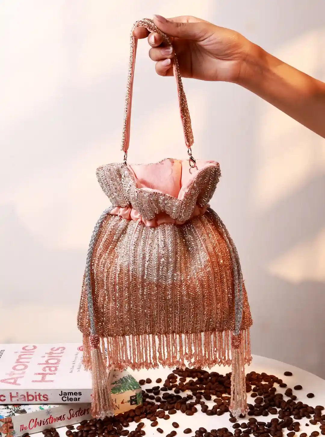 Handbag with fringe design held by a hand, placed on coffee beans with a book in the background.