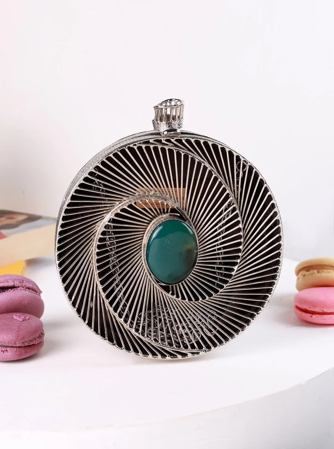 Circular metallic object with a green gemstone on a white surface with macarons.