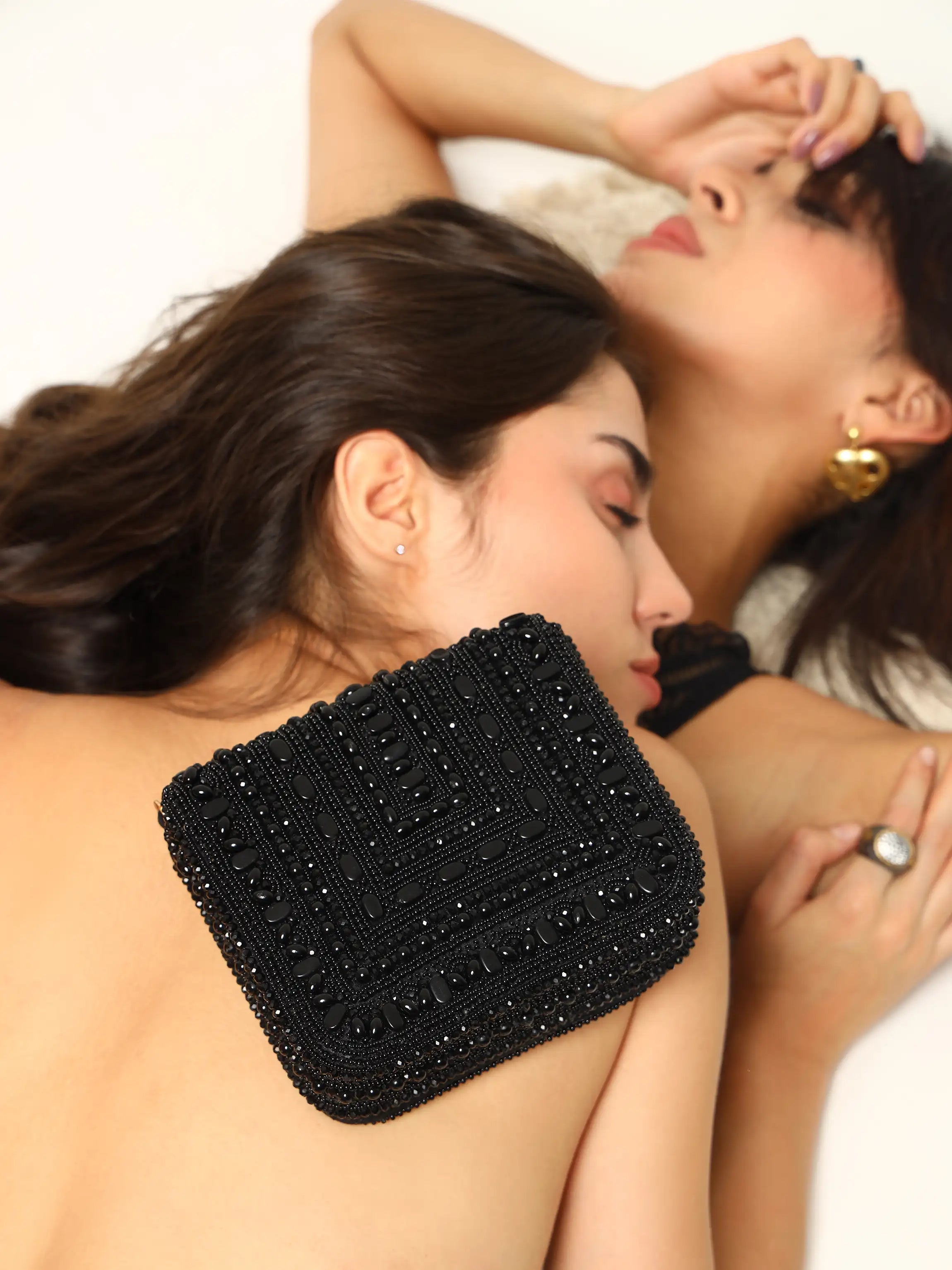 Two women lying down with a black beaded clutch bag on a white background
