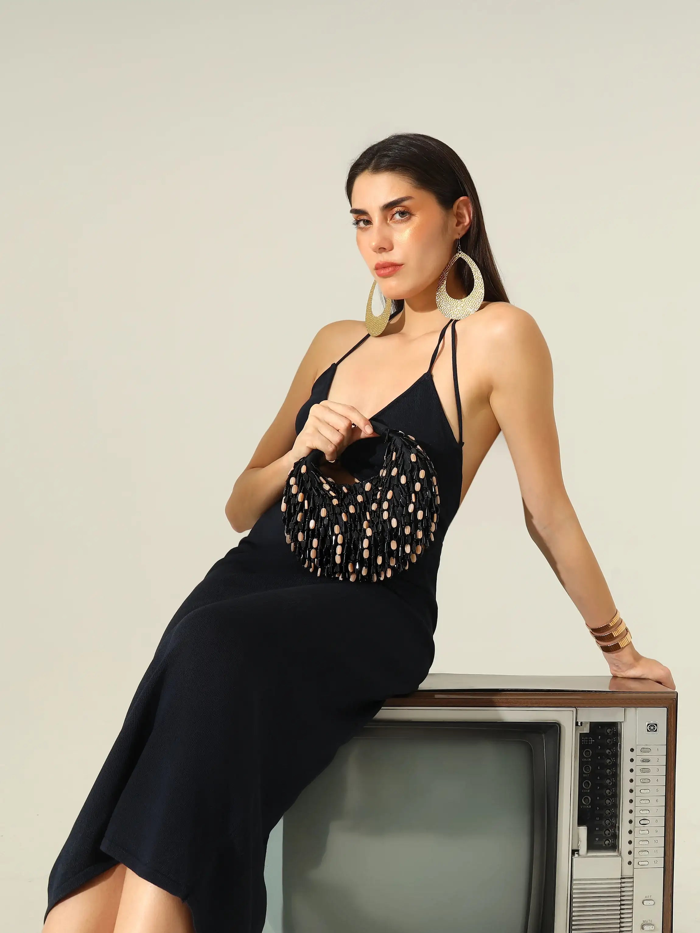 Woman in a black dress holding a patterned handbag, sitting on an old television set against a plain background.