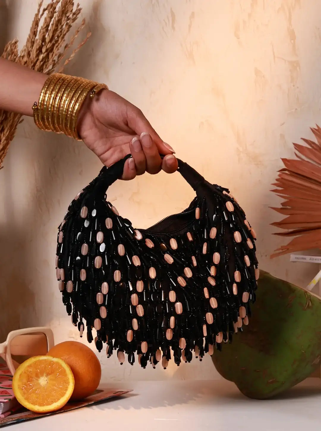 Black beaded handbag held by a hand with gold bangles, on a neutral background.