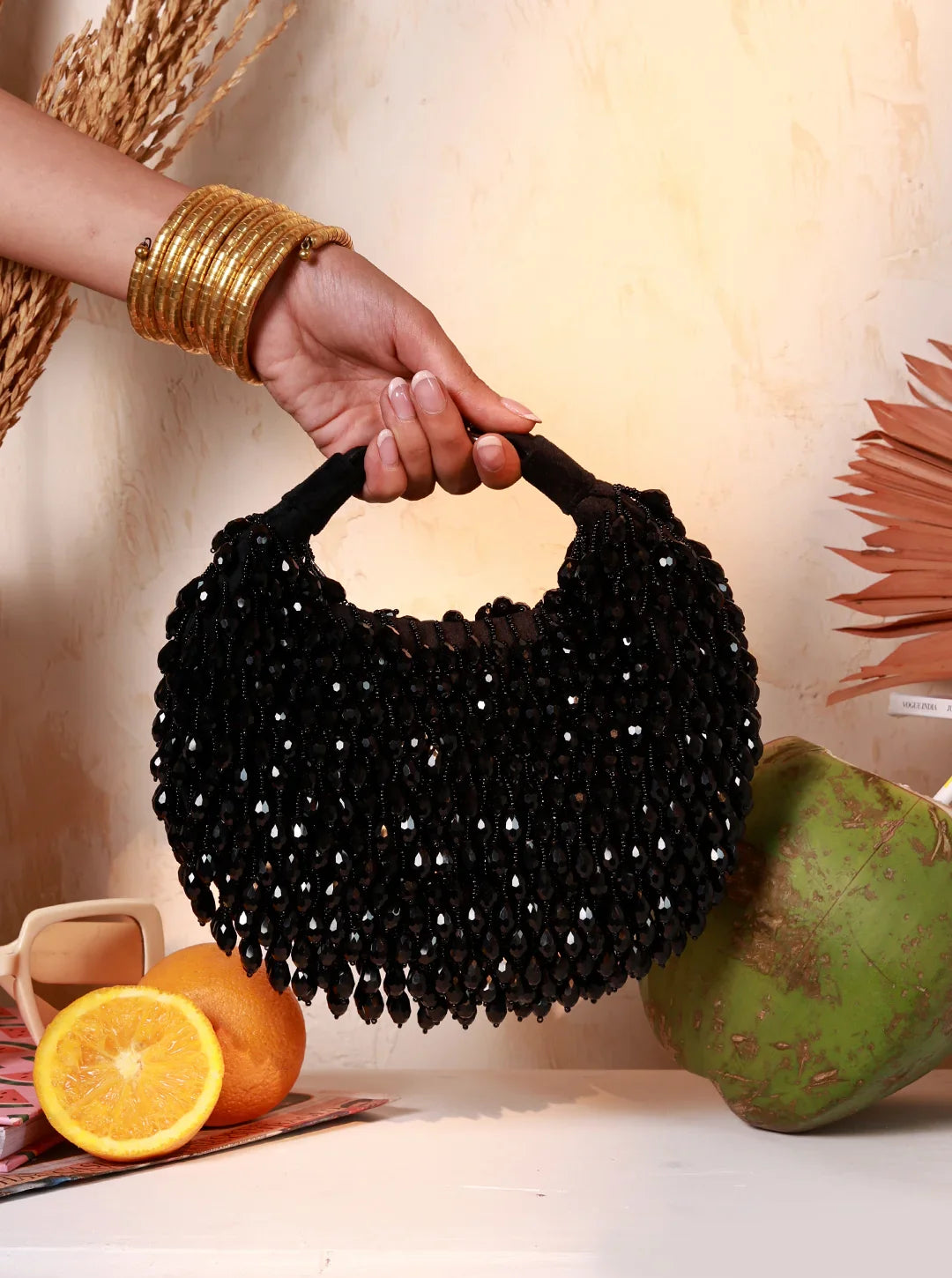Black beaded handbag held by a hand with gold bangles, on a neutral background with fruits.