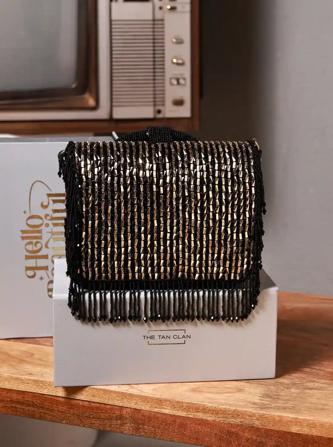 Black beaded clutch bag on a white box with 'The Tan Clan' branding, placed on a wooden surface.