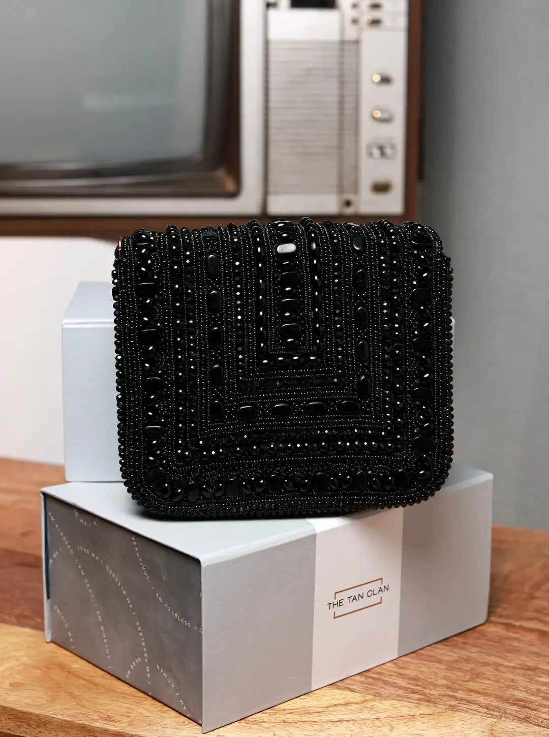 Black beaded clutch on a white box with 'The Tan Clan' branding, against a neutral background.
