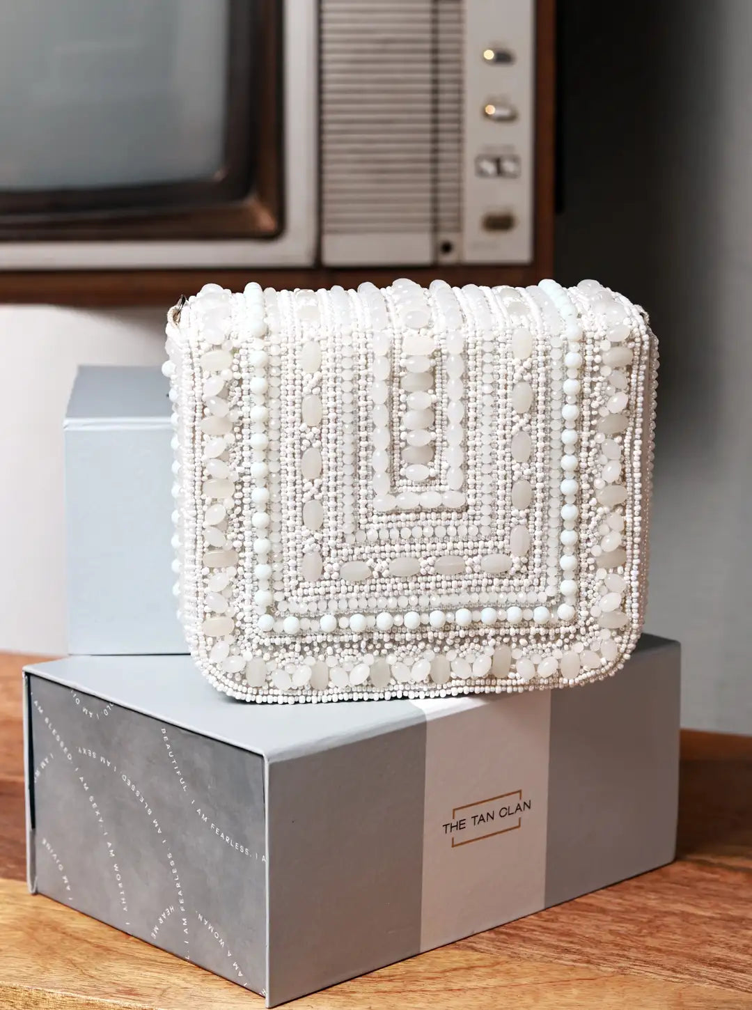 White beaded handbag on a box with 'The Tan Clan' branding, vintage television in the background