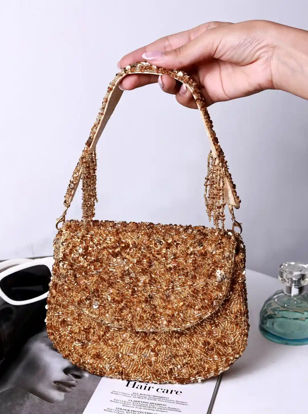 Gold beaded handbag held by a hand on a white surface with a magazine and bottle in the background.