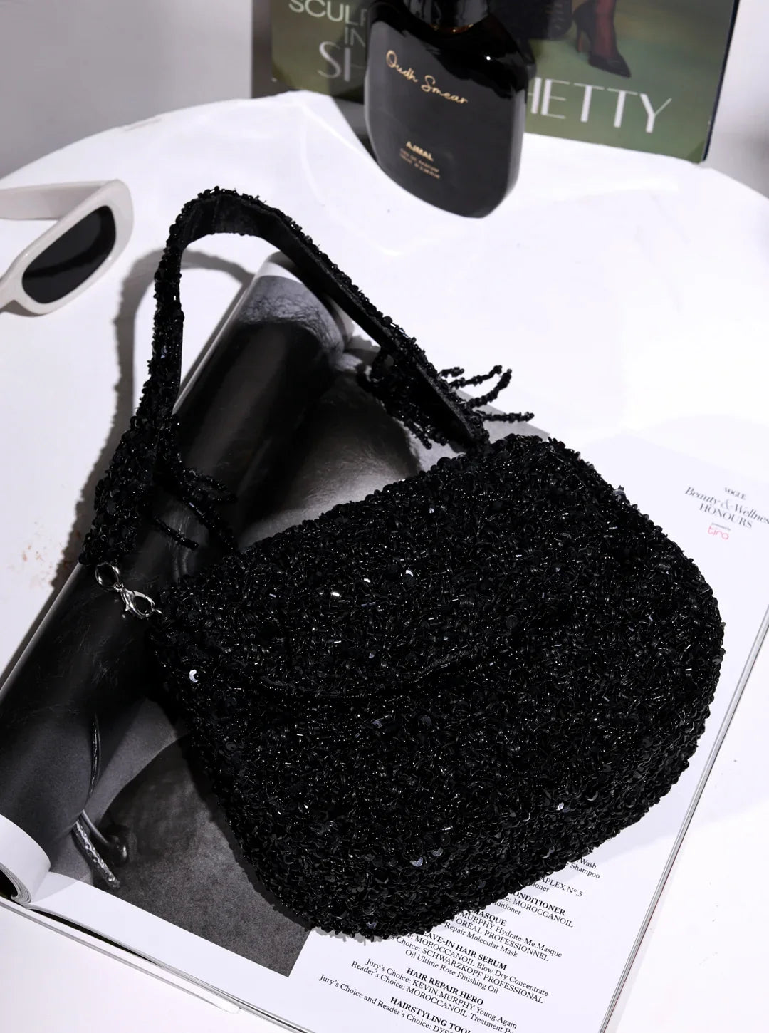 Black beaded handbag on a magazine with a bottle of perfume in the background
