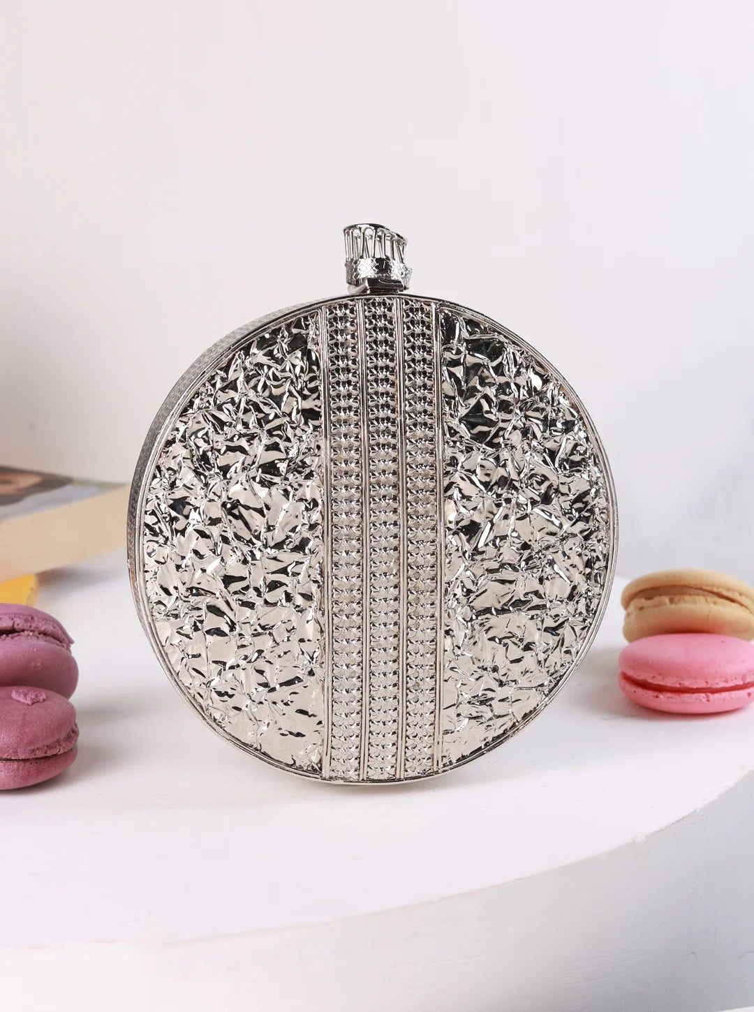 Silver round clutch with intricate design on a white surface with macarons.