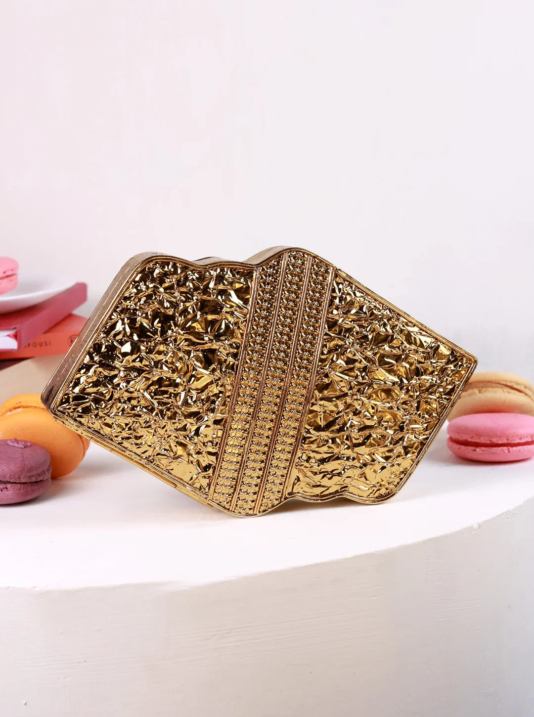 Gold clutch bag with intricate design on a white surface with macarons.
