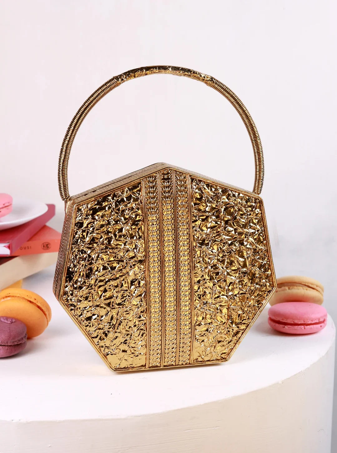 Gold hexagonal handbag with intricate patterns on a white surface with colorful macarons.