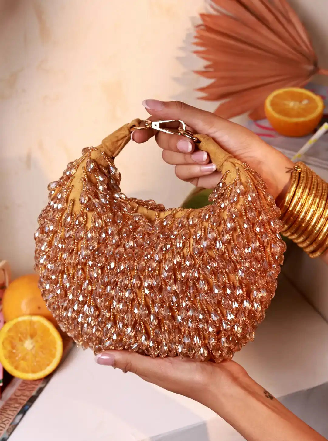 Hand holding a sparkling gold handbag with oranges and a fan in the background